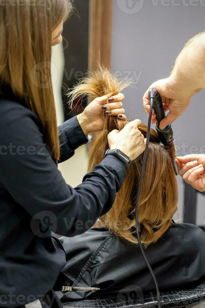 The rear view of two hairdressers are curling hair for a young woman with electric hair iron in a beauty salon. photo