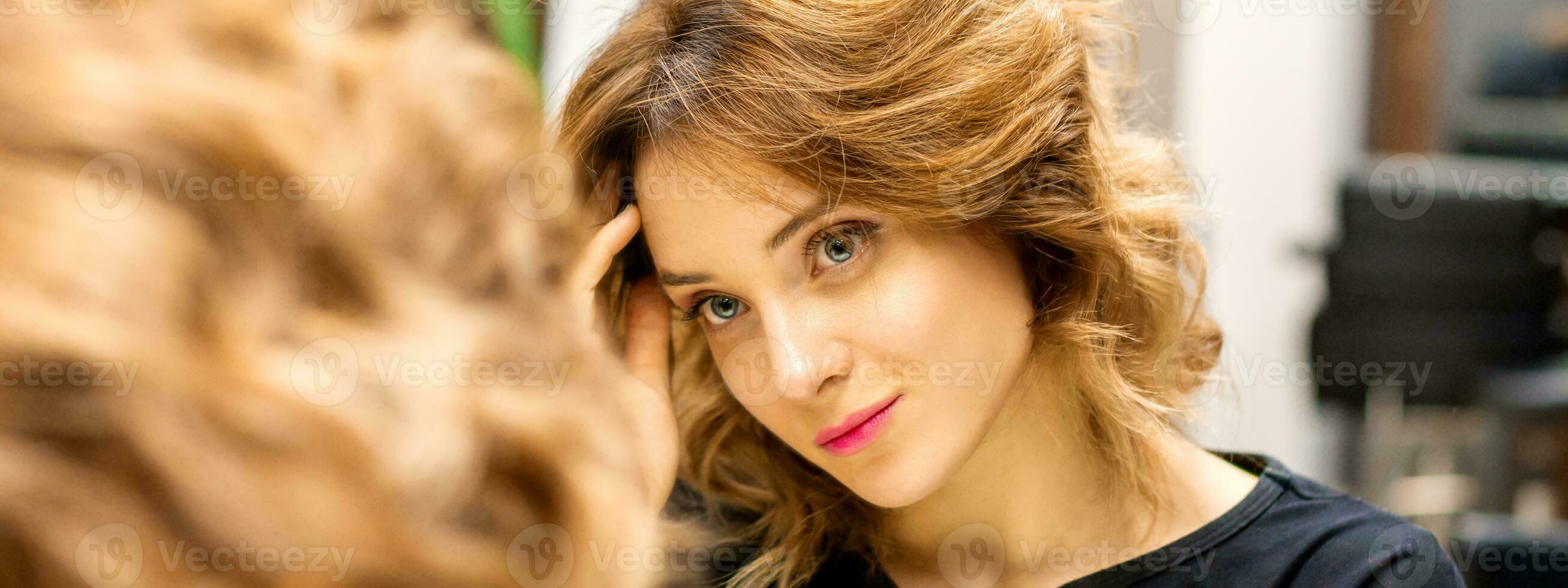 The beautiful young caucasian woman looks at her reflection in the mirror checking hairstyle and makeup in a beauty salon. photo