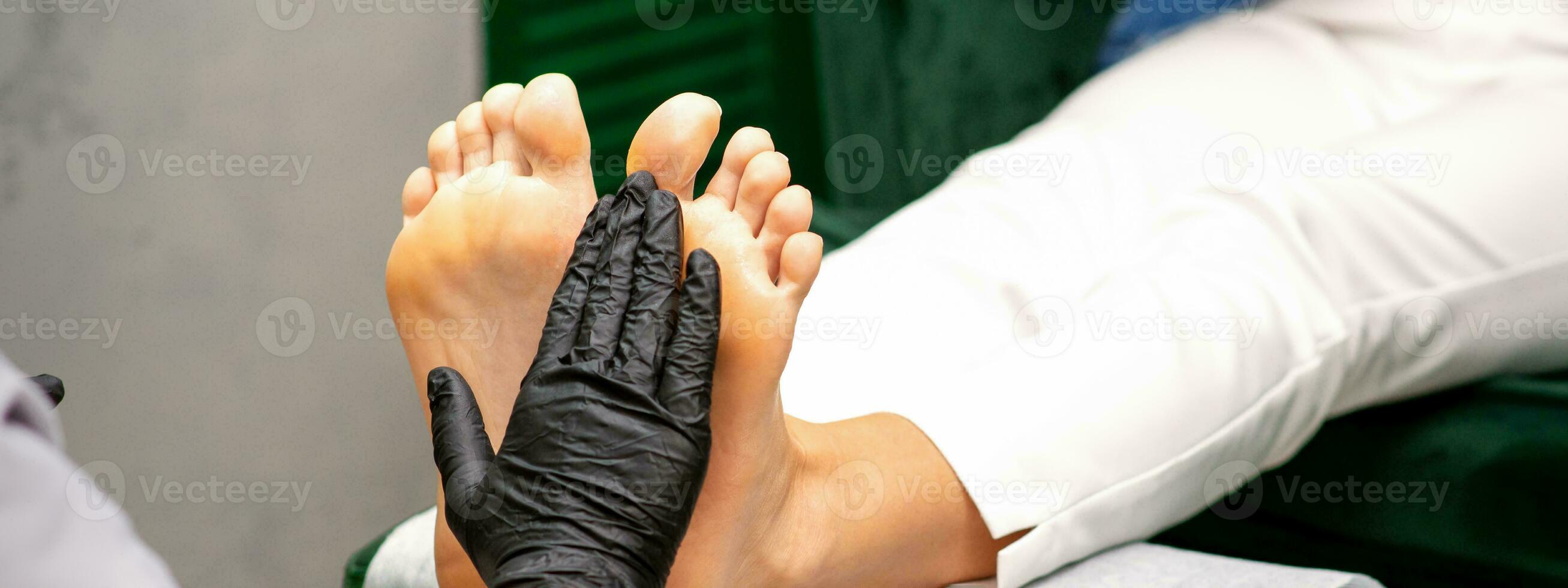 Moisturizing the feet. Hands of pedicure master in black gloves care about female feet. Foot massage. Pedicure beauty salon concept. photo