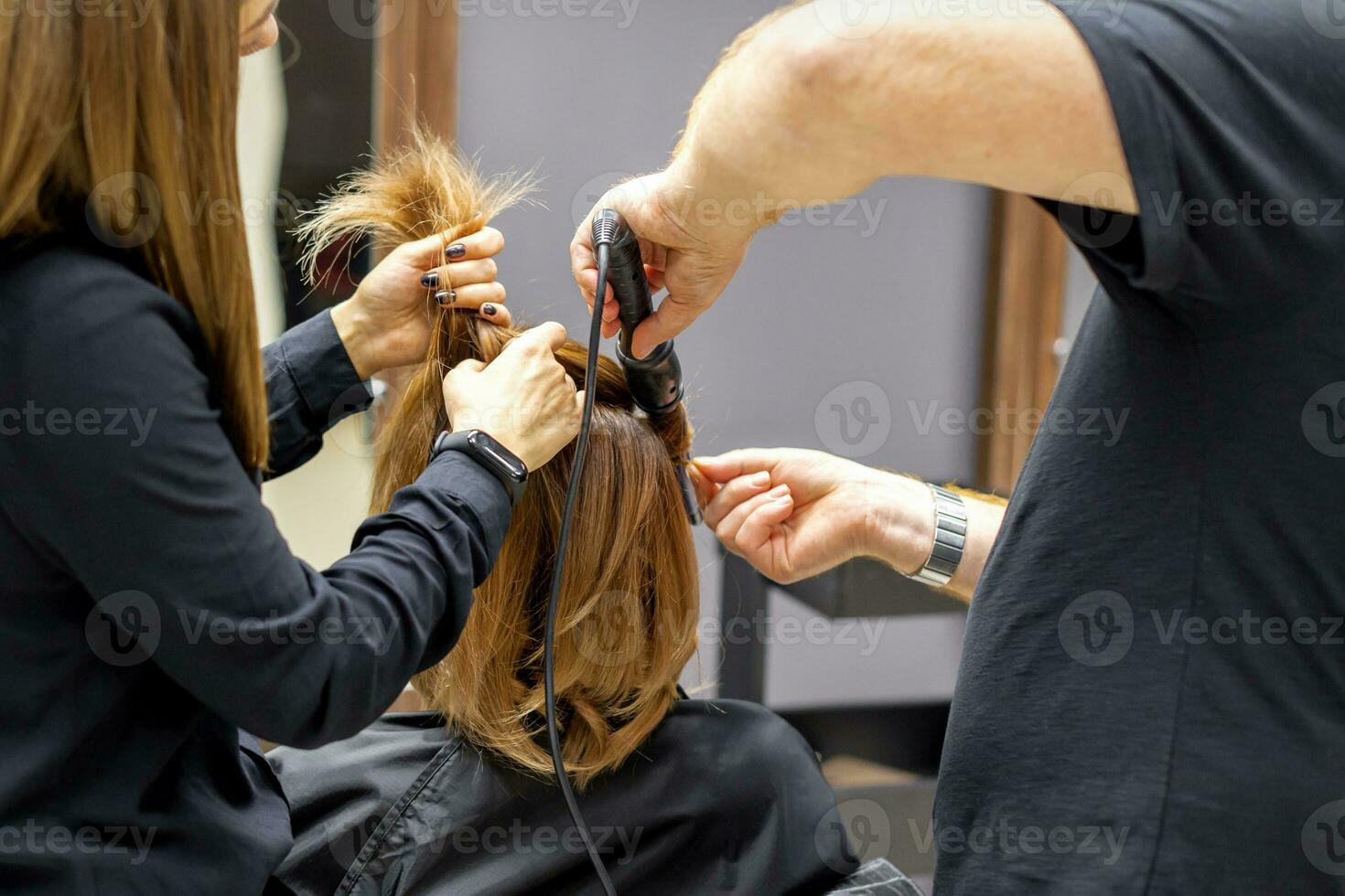 The rear view of two hairdressers are curling hair for a young woman with electric hair iron in a beauty salon. photo