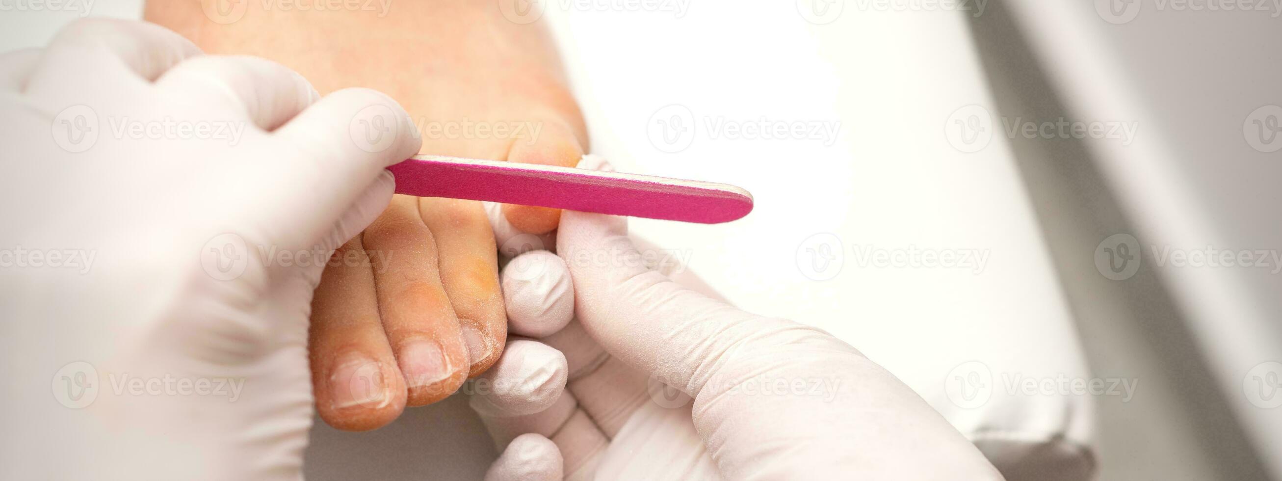 Pedicurist hands in protective rubber gloves filing toenails on feet with a nail file in a beauty salon. photo