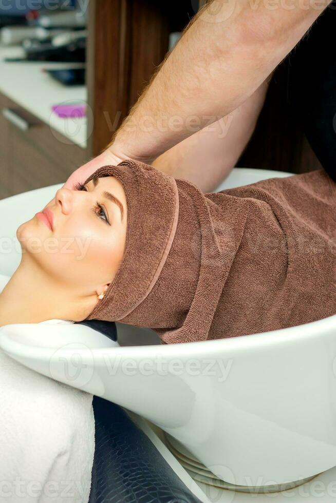 Gorgeous young relaxed woman getting her wet hair wrapped in a towel after washing by a professional hairdresser at the beauty salon. photo