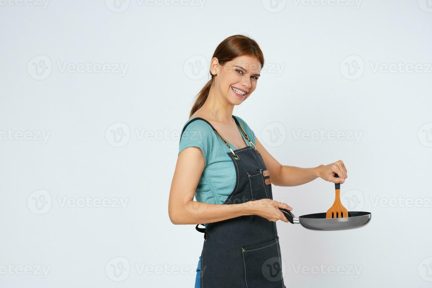 Young woman wearing kitchen apron cooking and holding pan and spatula isolated on white background. photo