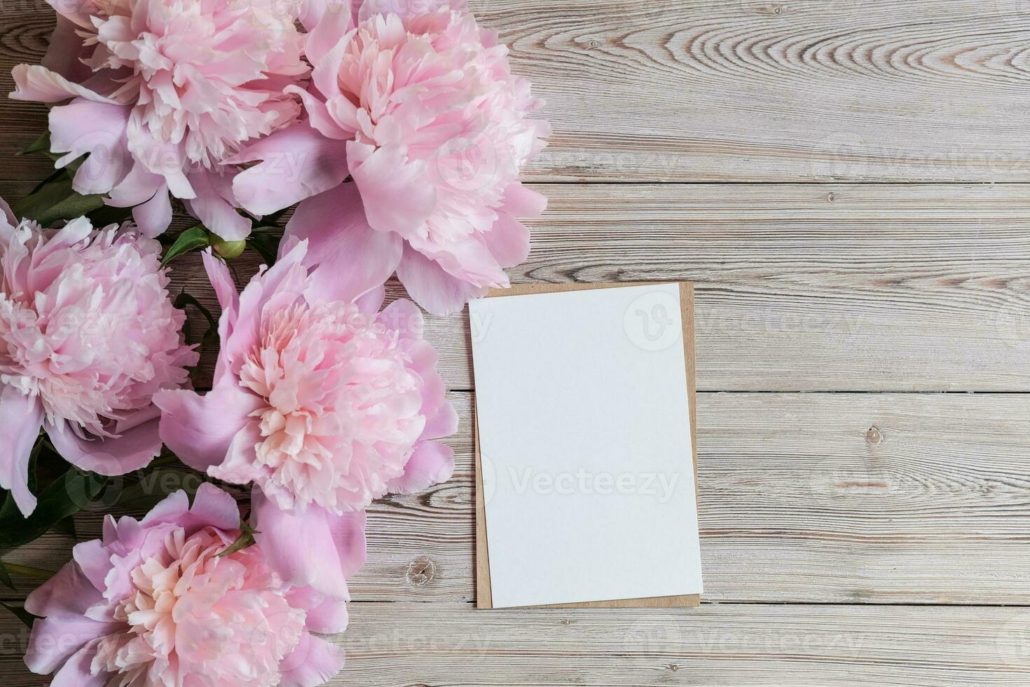 simula una tarjeta de papel blanco en blanco para texto y delicadas peonías sobre un fondo de madera. endecha plana, vista superior. lugar para el texto. tarjeta de felicitación foto