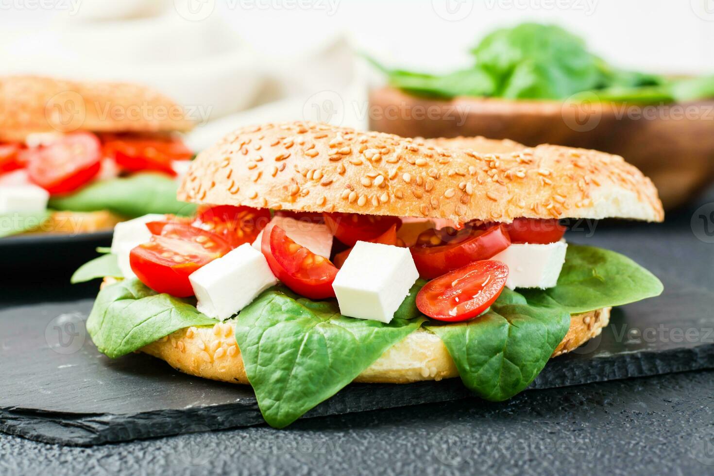 Delicious ready-to-eat bagel stuffed with tomatoes, feta and spinach leaves on a black background. Light healthy snack. Close-up photo