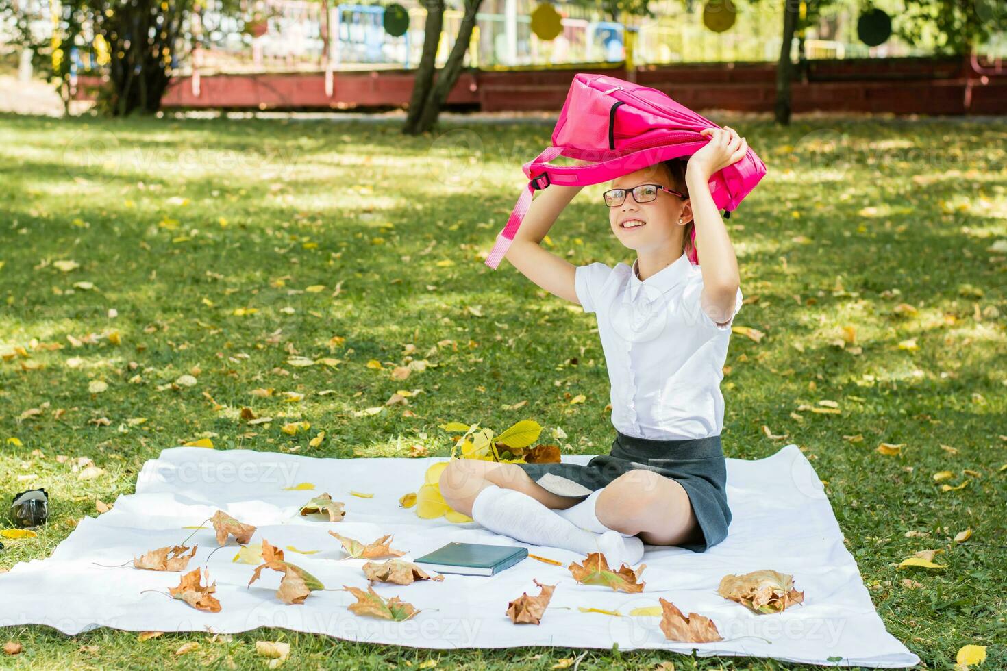 A cute schoolgirl with glasses smiles and holds a backpack above her head while sitting on a blanket in a sunny autumn park. Back to school concept photo