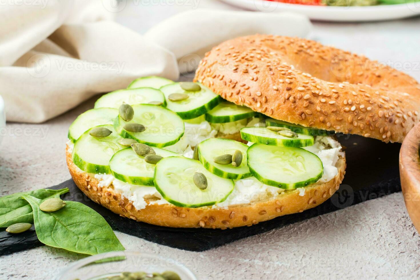 Ready-to-eat bagel stuffed with feta, cucumber and pumpkin seeds and spinach leaves on a slate board. Light Vitamin Healthy Snack. Close-up photo