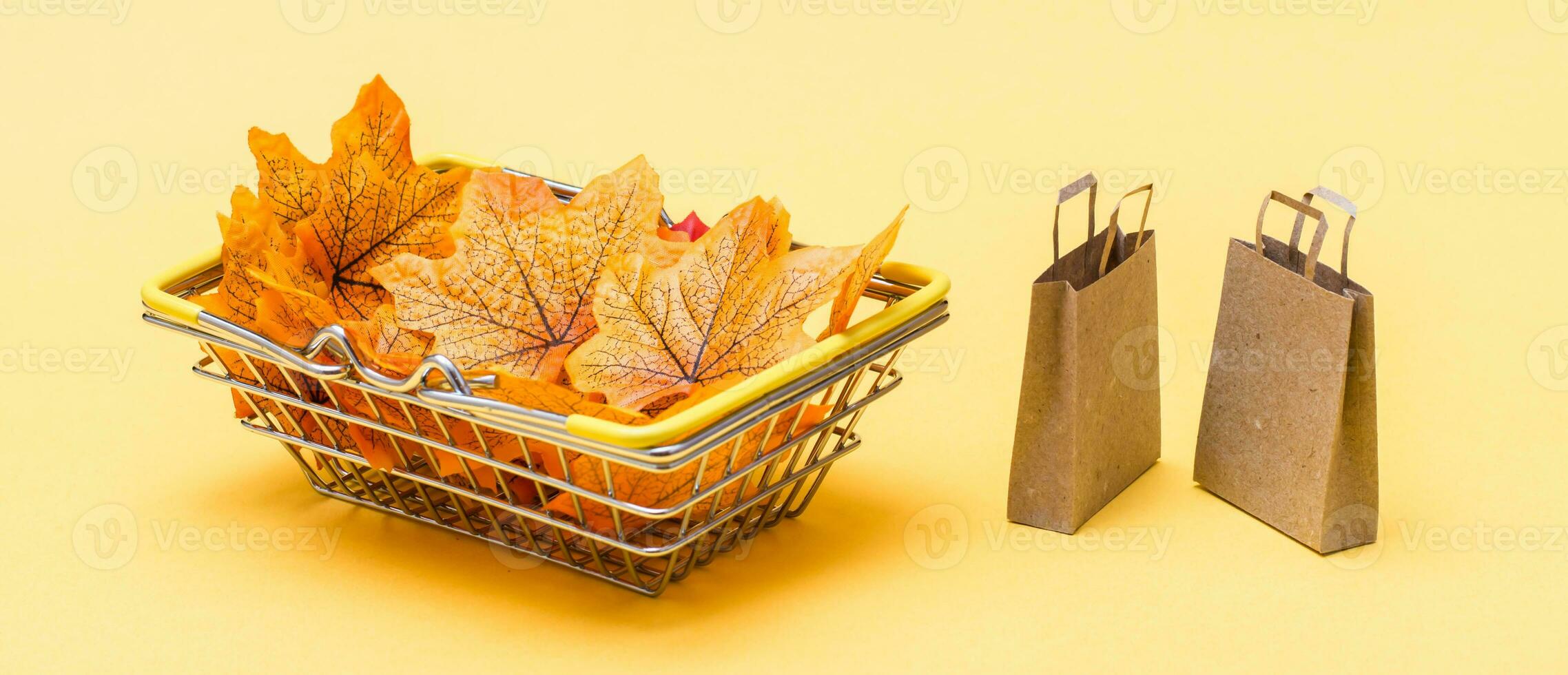 A metal shopping basket in a store filled with autumn leaves and a couple of craft paper bags on a yellow background. Black friday gift sales. Web banner photo