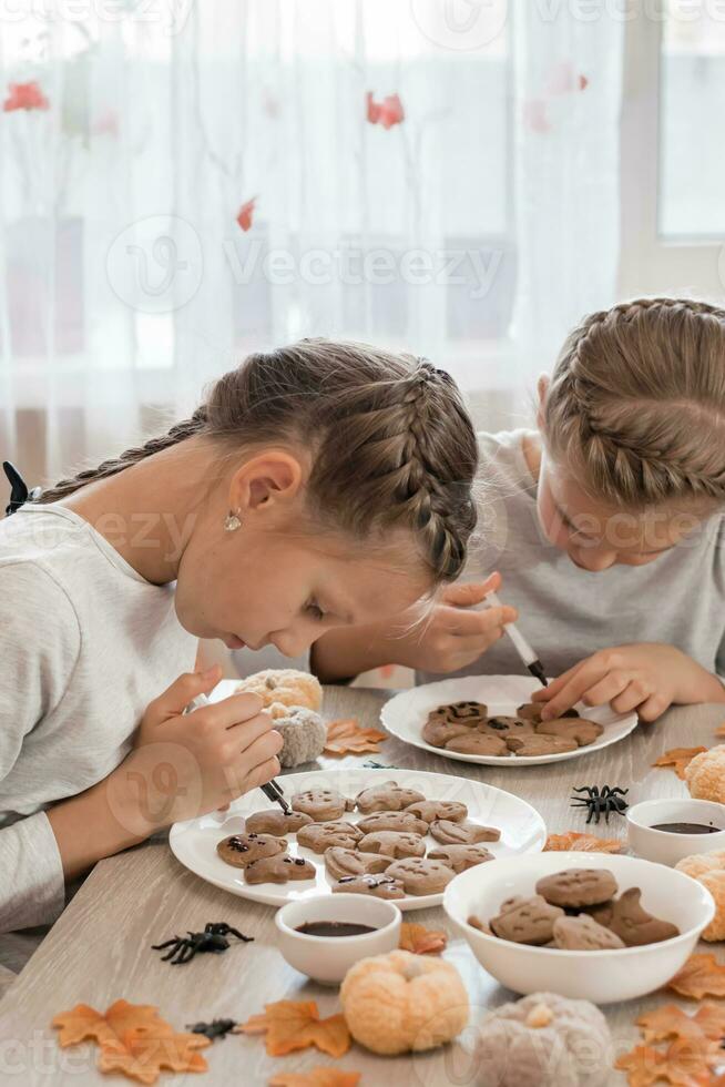 Preparing to celebrate halloween and preparing a treat. Two girls decorate halloween gingerbread cookies on plates with chocolate icing. Lifestyle photo