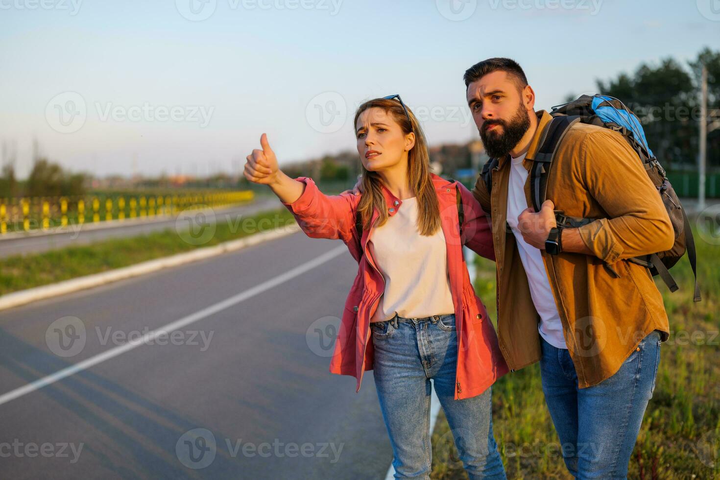 Adult couple hitchhiking on roadside trying to stop car. They are anxious and bored. photo