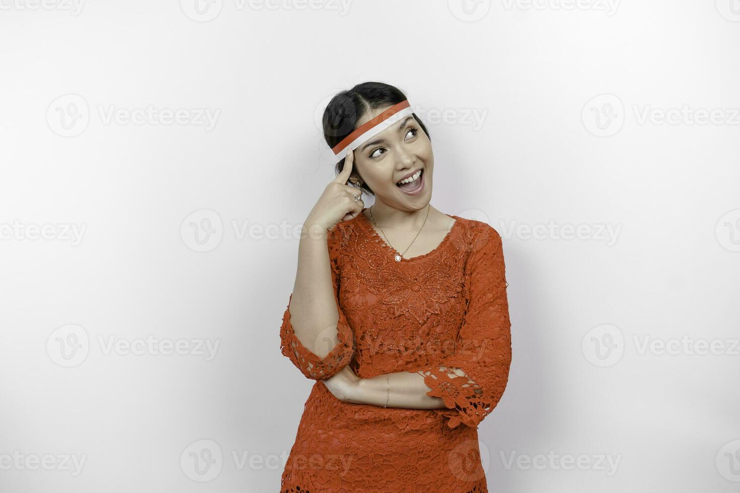 A smart young woman wearing headband and red kebaya is looking aside pointing to an idea on copy space , isolated by white background. Indonesia's independence day. photo