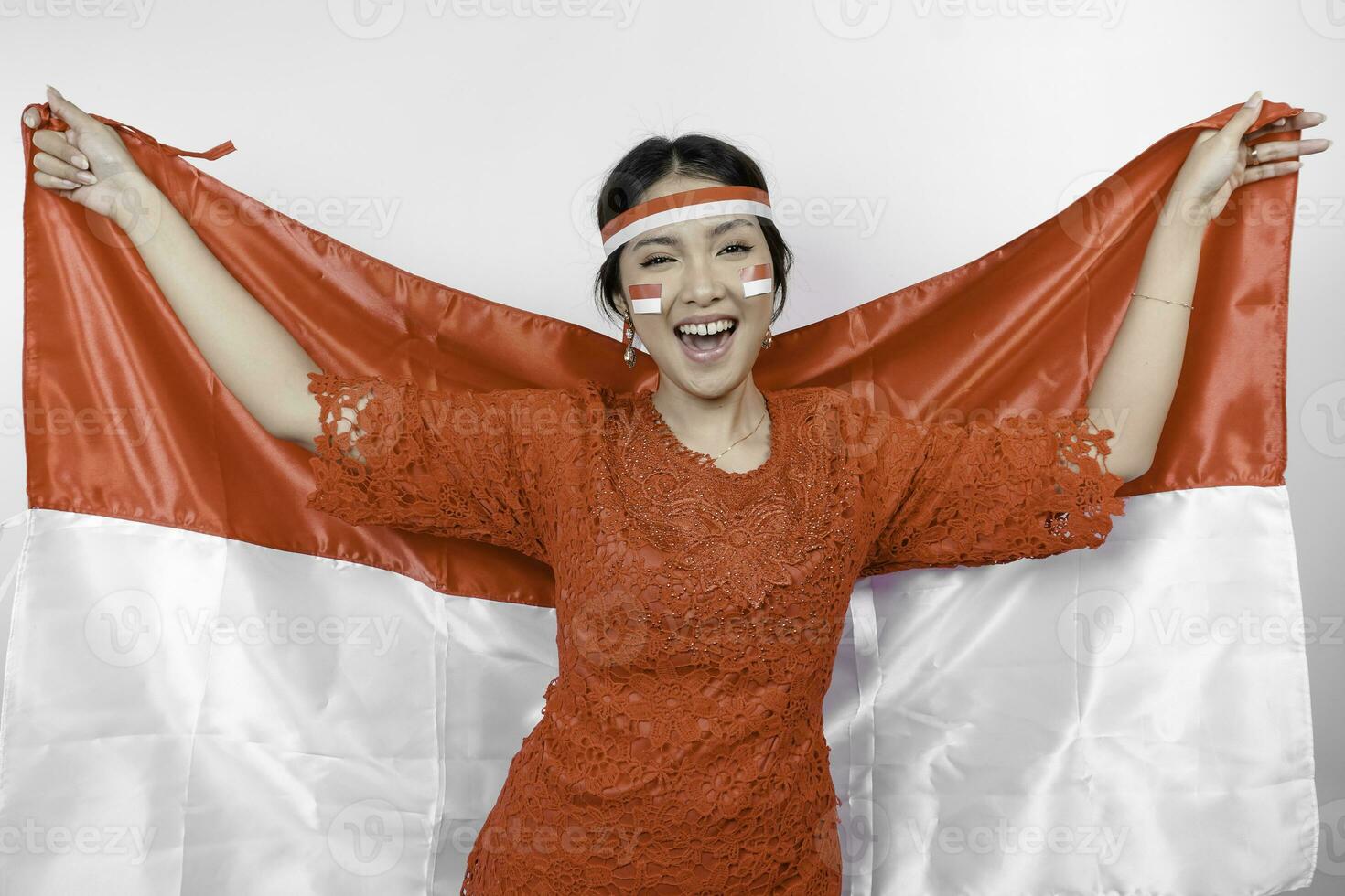 Happy smiling Indonesian woman wearing red kebaya and headband holding Indonesia's flag to celebrate Indonesia Independence Day isolated over white background. photo