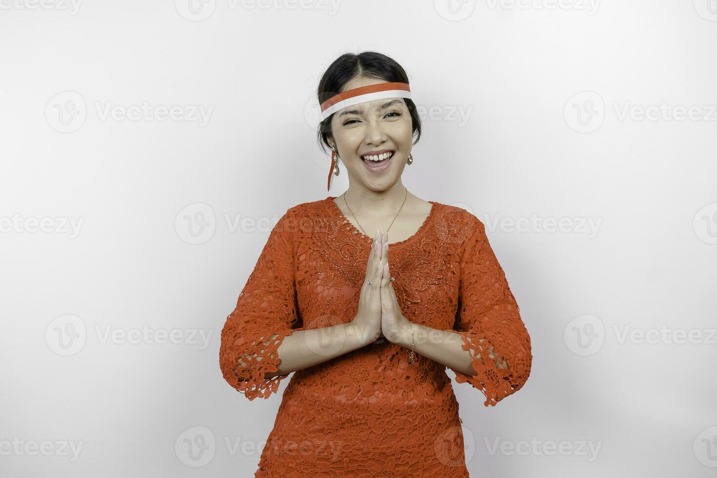 A friendly Indonesian woman is wearing red kebaya gesturing traditional greeting and Indonesia's flag headband to celebrate Indonesia Independence Day. Isolated by white background. photo