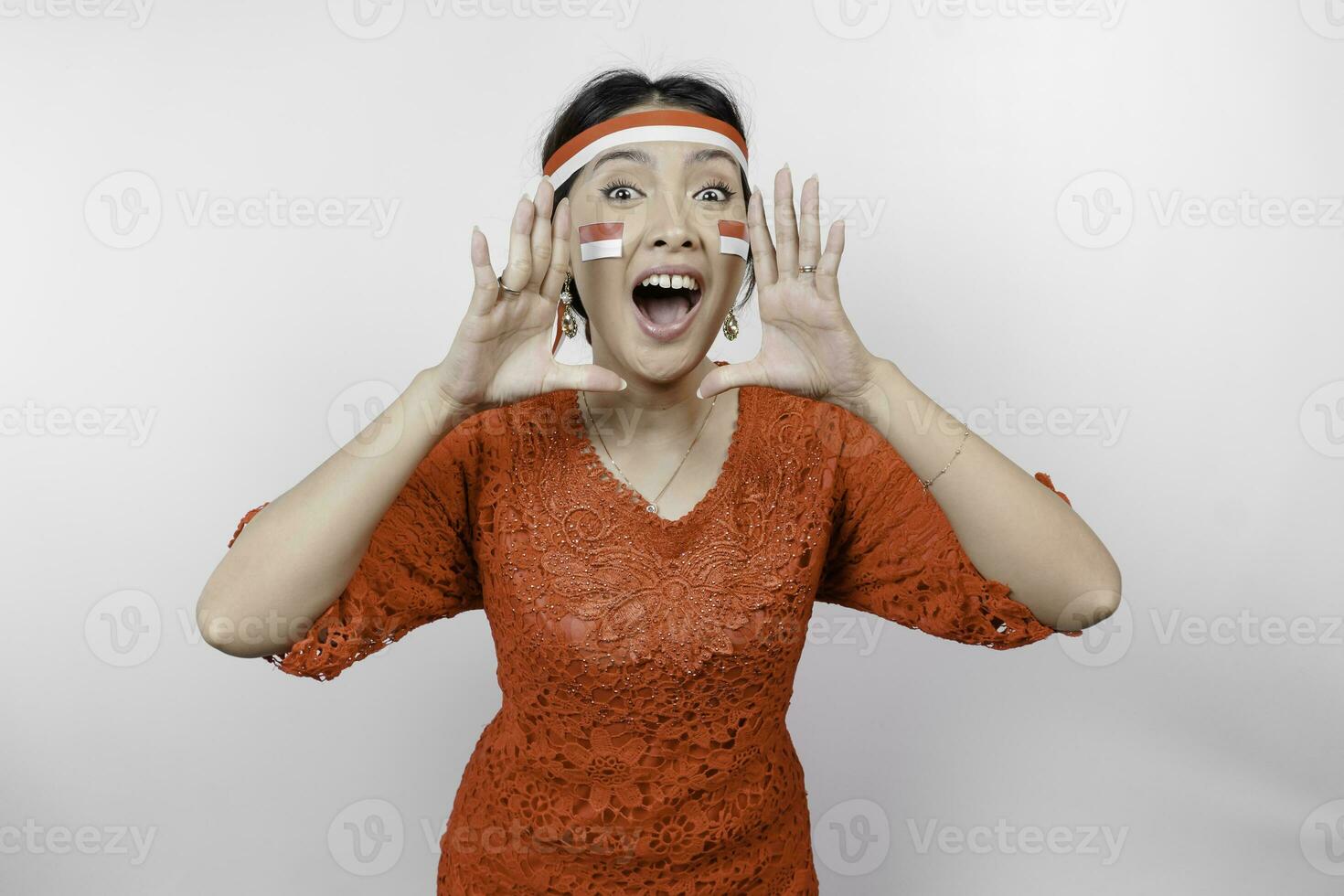 Young beautiful woman wearing a red kebaya and headband is shouting and screaming loud with a hand on her mouth. Indonesia's independence day concept. photo