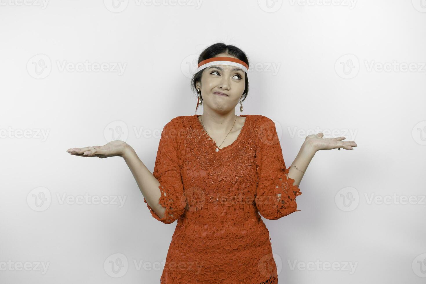 A thoughtful young woman wearing headband and red kebaya is looking aside confuse between choices, isolated by white background. Indonesia's independence day. photo