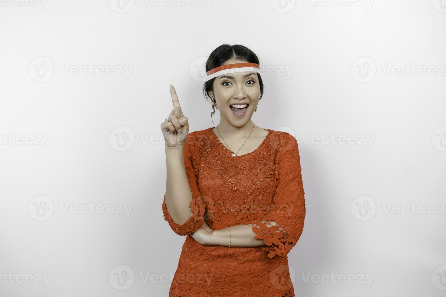 Excited Asian woman wearing red kebaya and flag headband, pointing at the copy space on top of her, isolated by white background photo