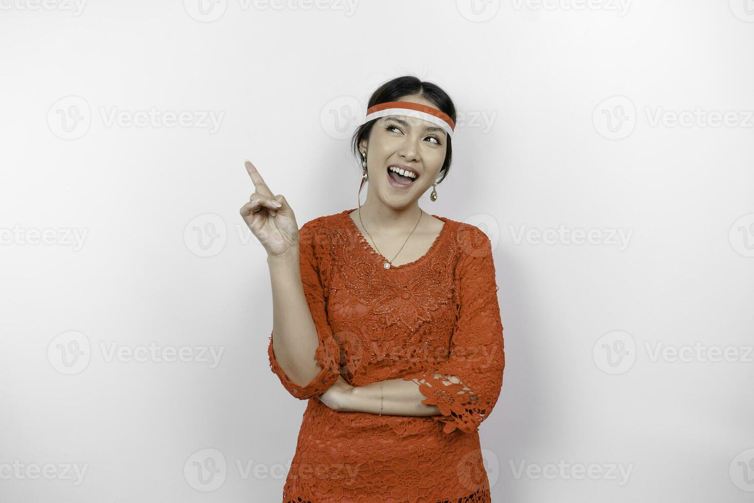 Excited Asian woman wearing red kebaya and flag headband, pointing at the copy space on top of her, isolated by white background photo