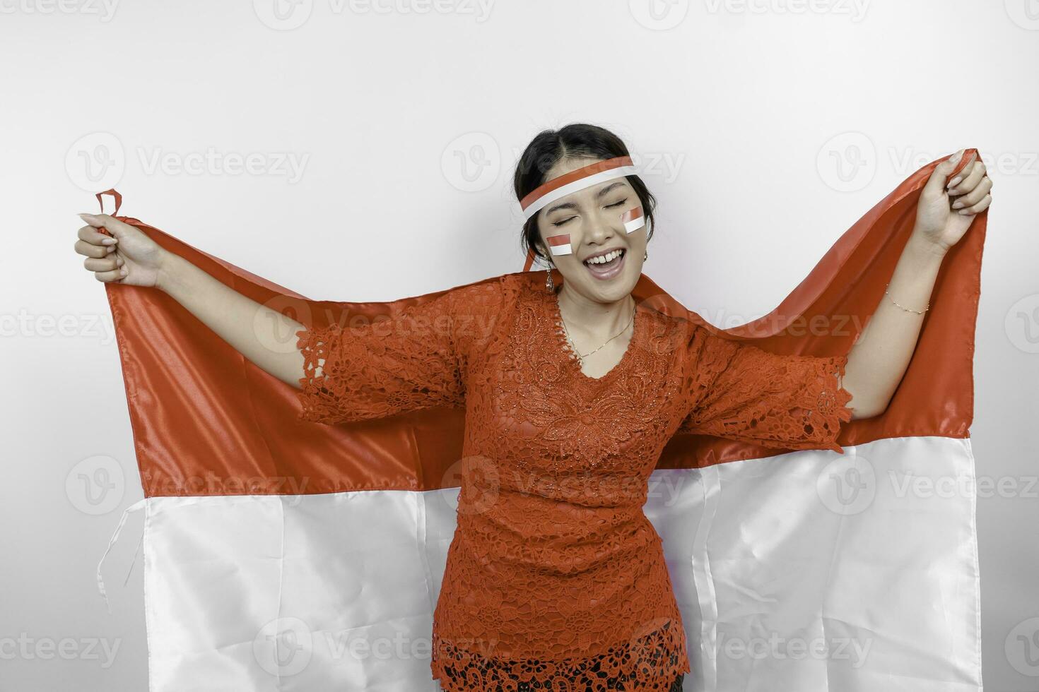 Happy smiling Indonesian woman wearing red kebaya and headband holding Indonesia's flag to celebrate Indonesia Independence Day isolated over white background. photo