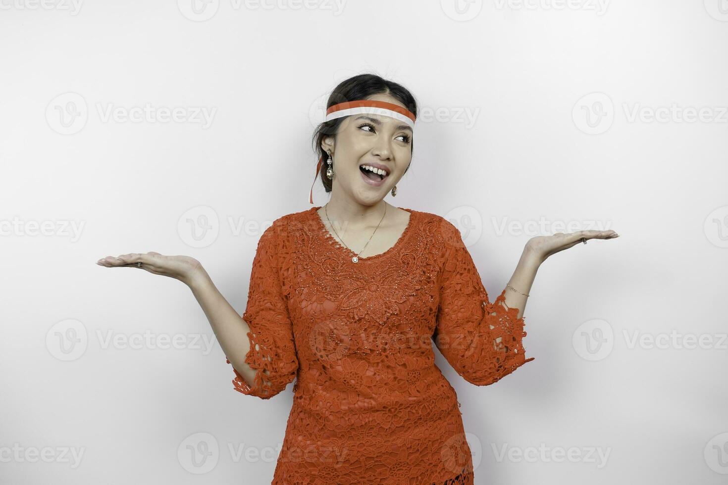 Excited Asian woman wearing red kebaya and headband, pointing at the copy space beside her, isolated by white background. Indonesia's independence day concept. photo