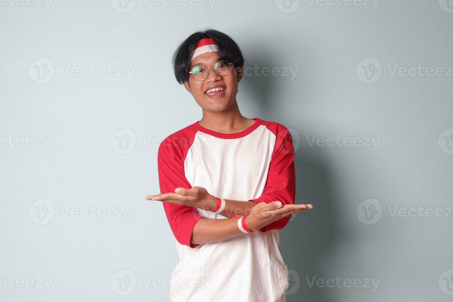 Portrait of attractive Asian man in t-shirt with red and white ribbon on head, choosing between 2 different options, holding two hands with empty space. Decision concept. Isolated image on gray photo