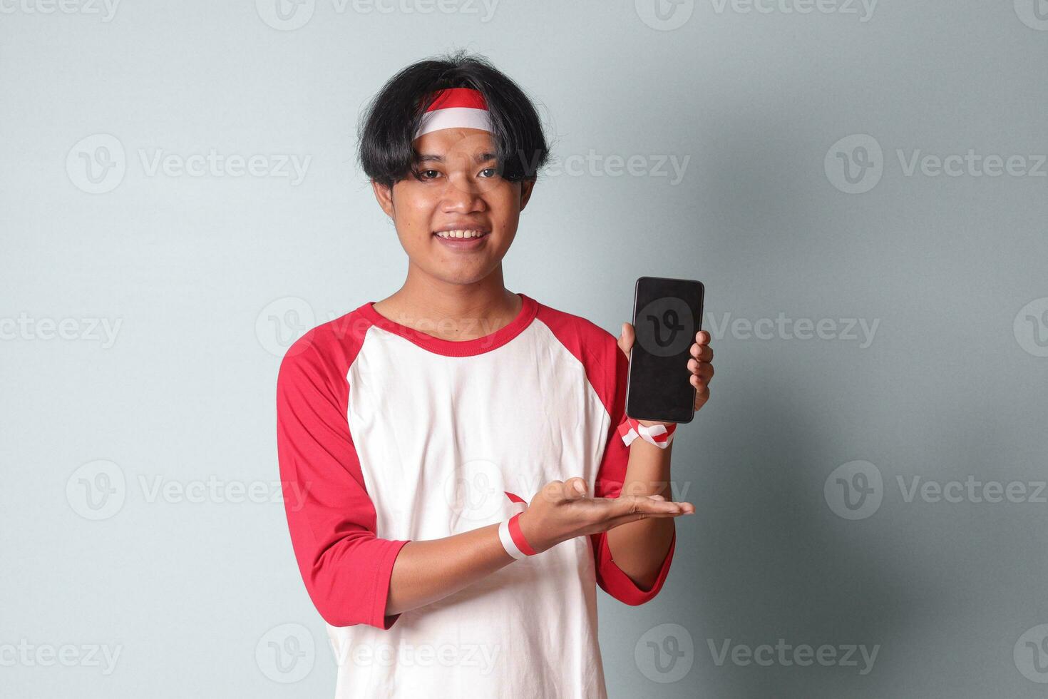 Portrait of attractive Asian man in t-shirt with red and white ribbon on head, holding and showing blank screen of mobile phone for mock-up. Isolated image on gray background photo