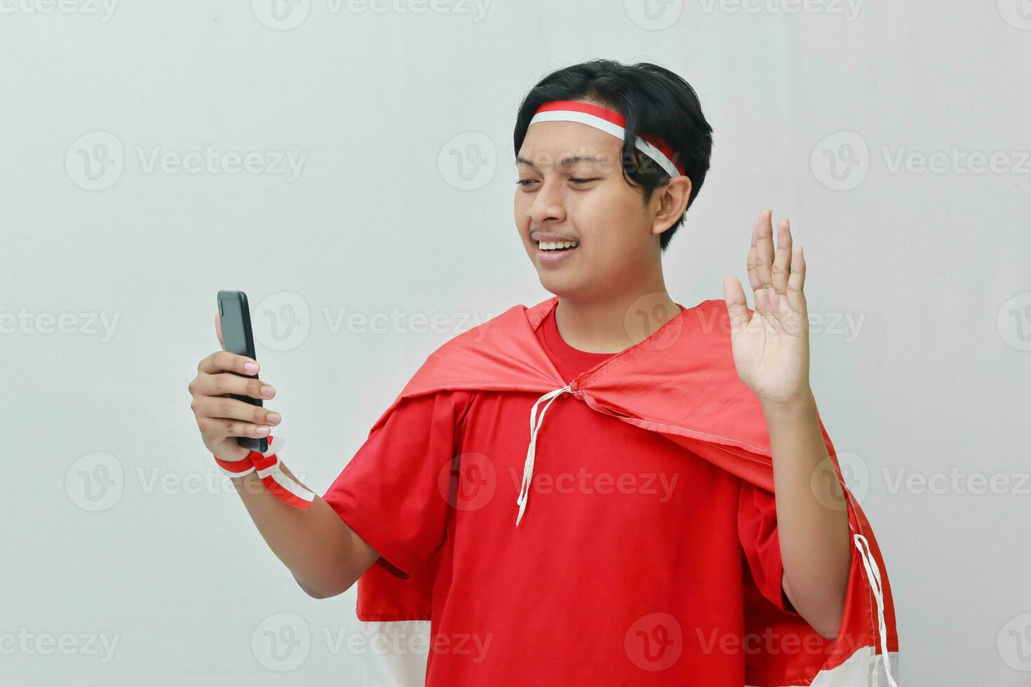 Portrait of attractive Asian man in t-shirt with red white ribbon on head with flag on his shoulder as a cloak, waving hand saying hi on his mobile phone during video calling. Isolated image on gray photo