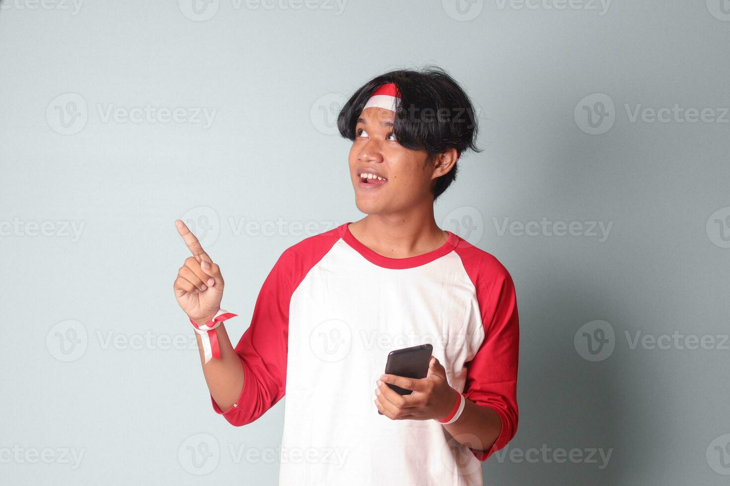Portrait of attractive Asian man in t-shirt with red and white ribbon on head, holding mobile phone while raising up Indonesia flag and pointing away. Isolated image on gray background photo