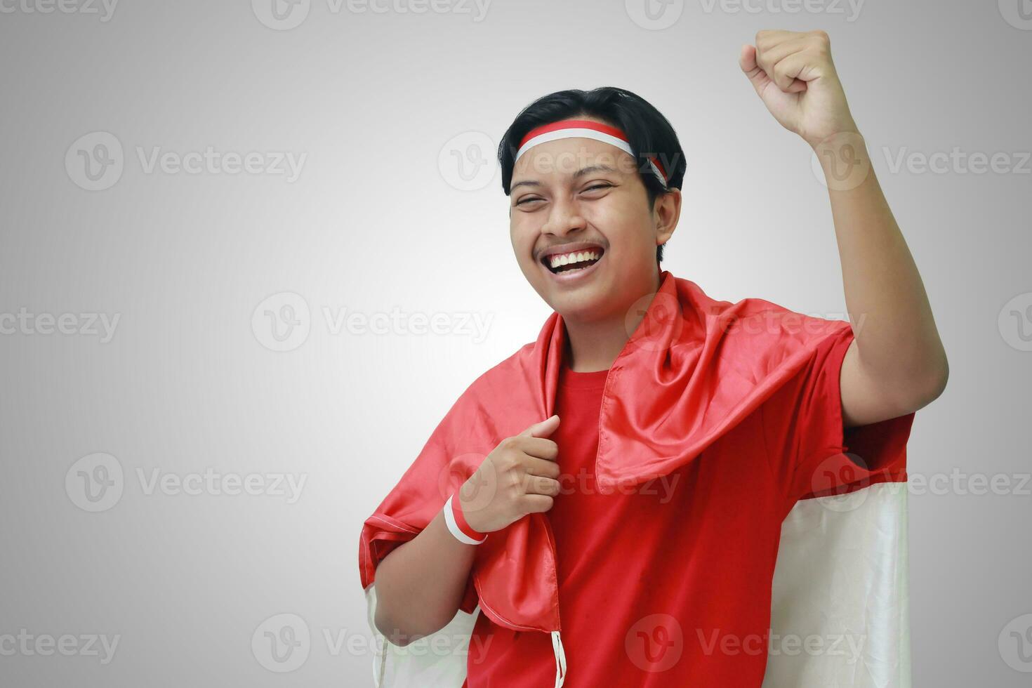 Portrait of attractive Asian man in t-shirt with red white ribbon on head with flag on his shoulder as a cloak, raising his fist, celebrating Indonesia's independence day. Isolated image on gray photo