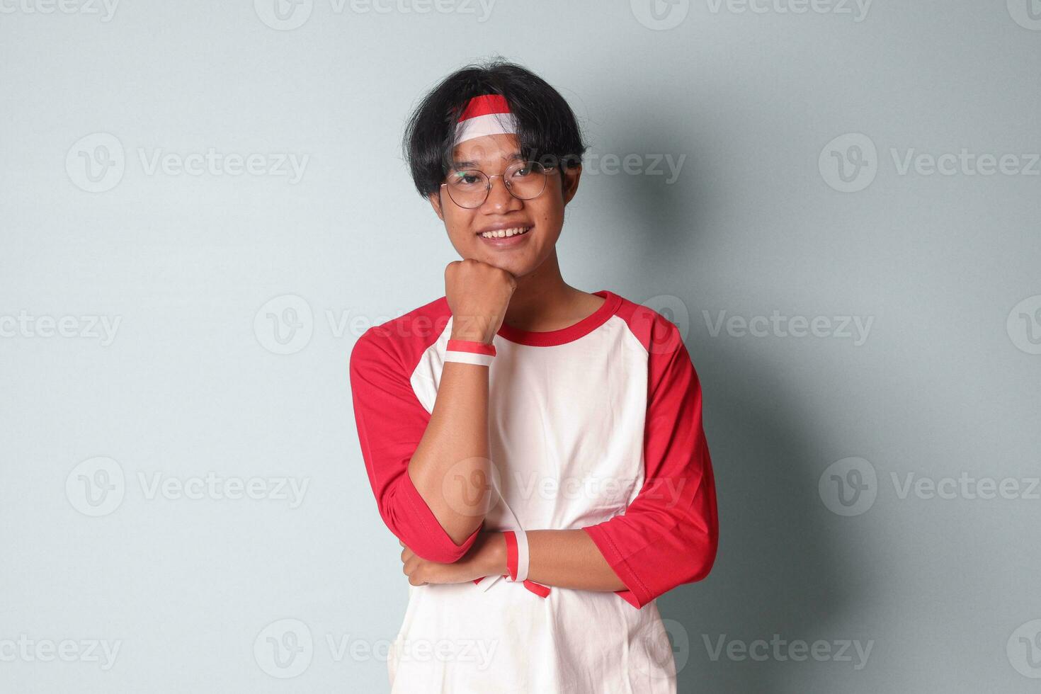 retrato de atractivo asiático hombre en camiseta con rojo y blanco cinta en cabeza, en pie en contra gris fondo, pensando acerca de pregunta con mano en barbilla foto