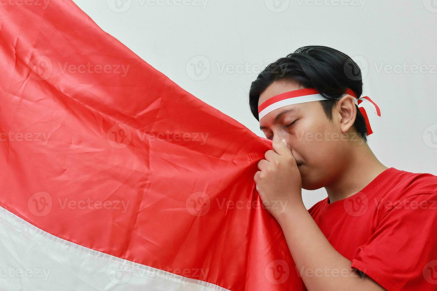 Portrait of respectful Asian man in t-shirt with headband ribbon on head, kissing red and white flag of Indonesia. Isolated image on gray background photo