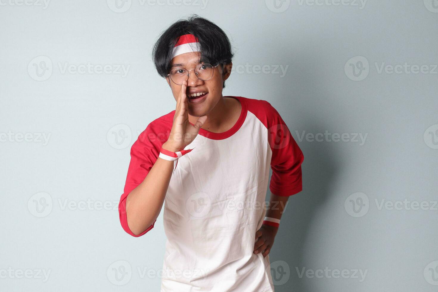 Portrait of attractive Asian man in t-shirt with red and white ribbon on head, whispering malicious talk conversation, hand on mouth telling secret rumor. Isolated image on gray background photo