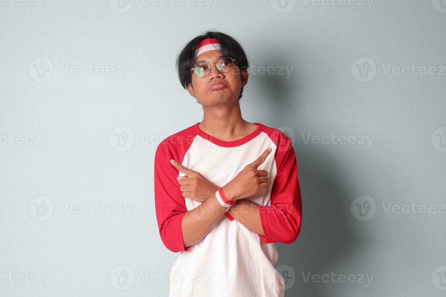 Portrait of attractive Asian man in t-shirt with red and white ribbon on head, choosing between 2 different options, holding two hands with empty space. Decision concept. Isolated image on gray photo