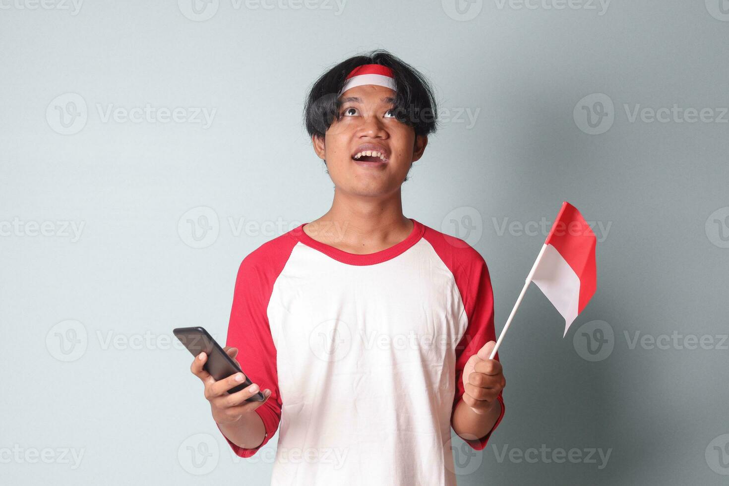 Portrait of attractive Asian man in t-shirt with red and white ribbon on head, holding mobile phone while raising up Indonesia flag. Isolated image on gray background photo