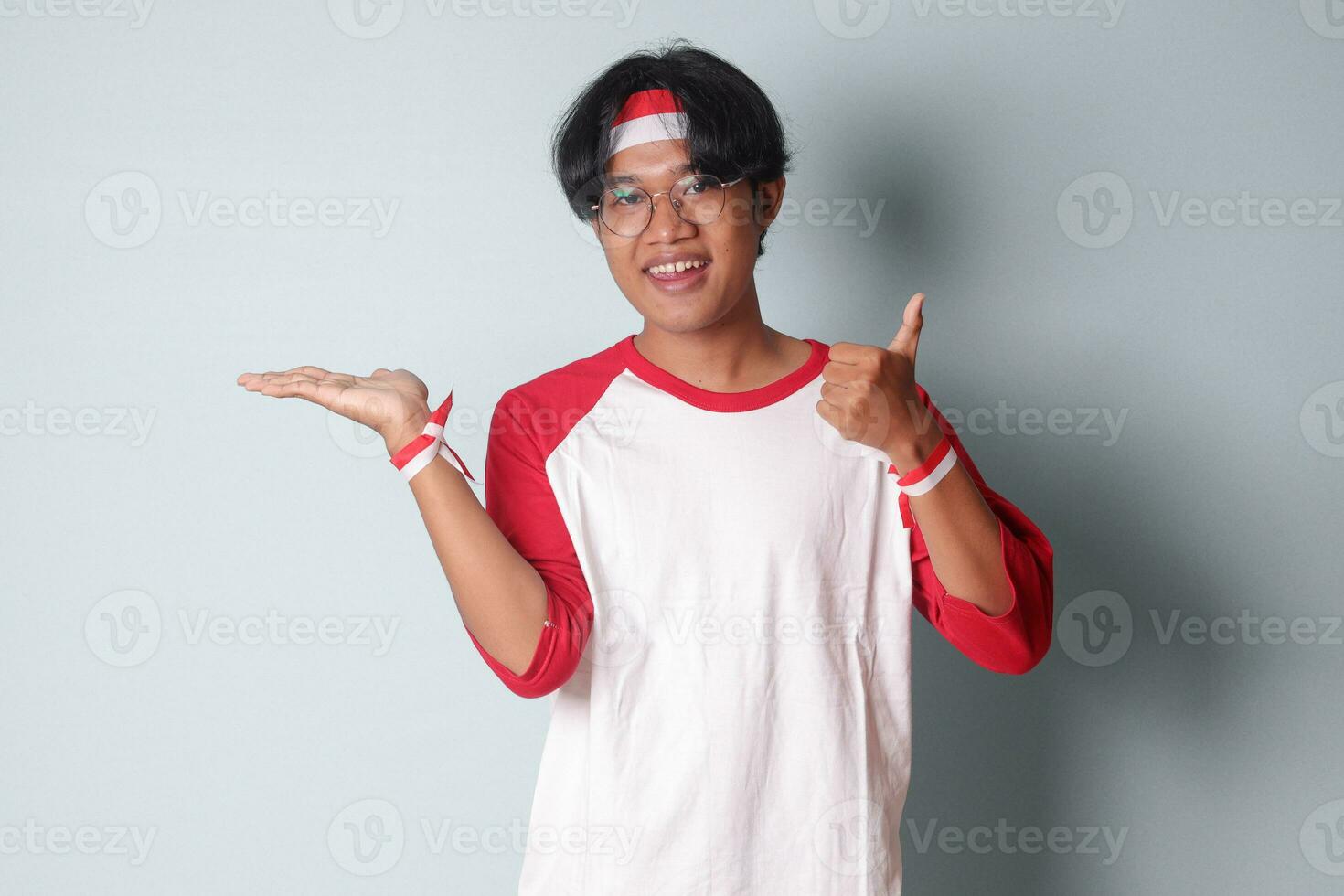 retrato de atractivo asiático hombre en camiseta con rojo y blanco cinta en cabeza, levantamiento su puño mientras demostrando producto. aislado imagen en gris antecedentes foto