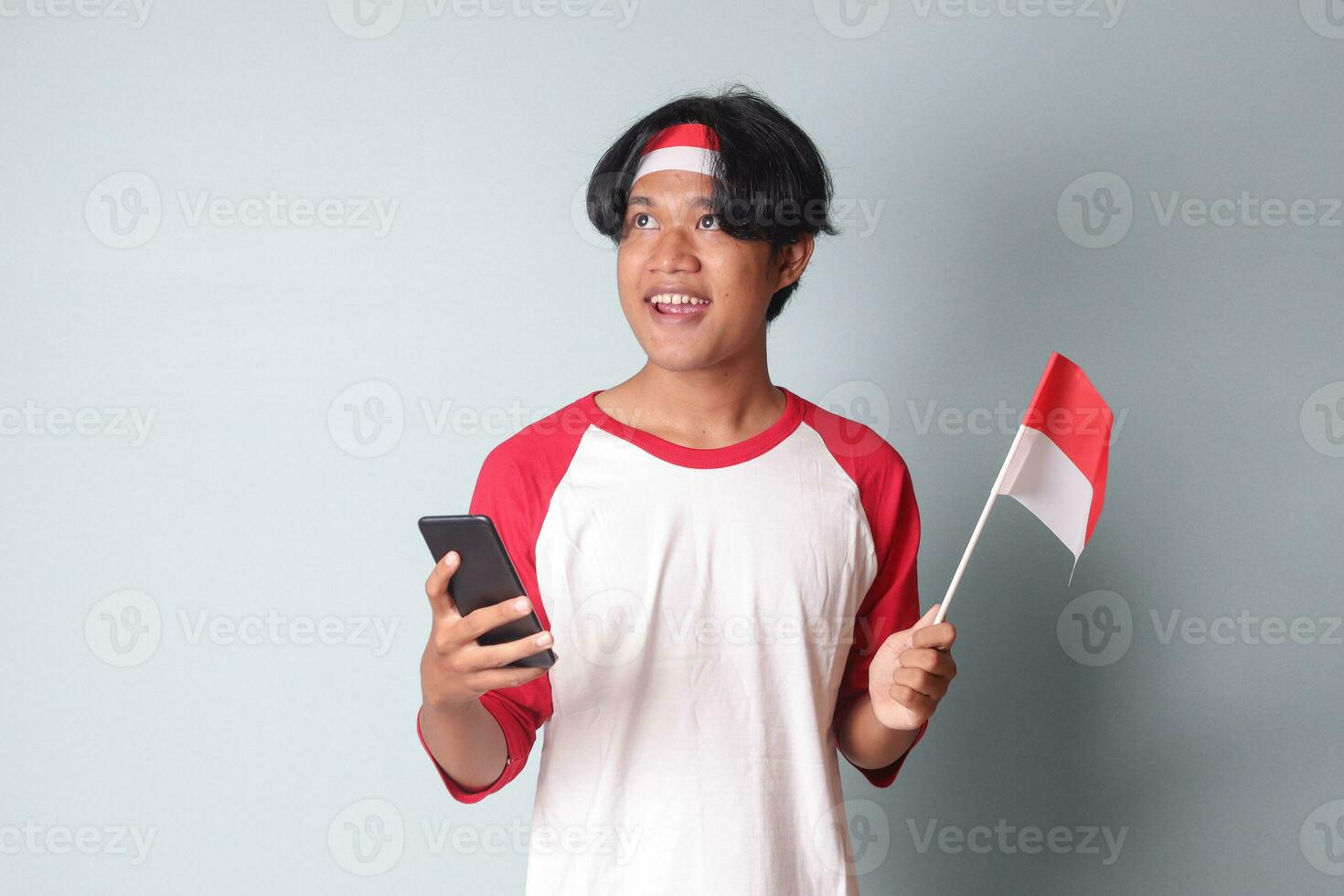 Portrait of attractive Asian man in t-shirt with red and white ribbon on head, holding mobile phone while raising up Indonesia flag. Isolated image on gray background photo