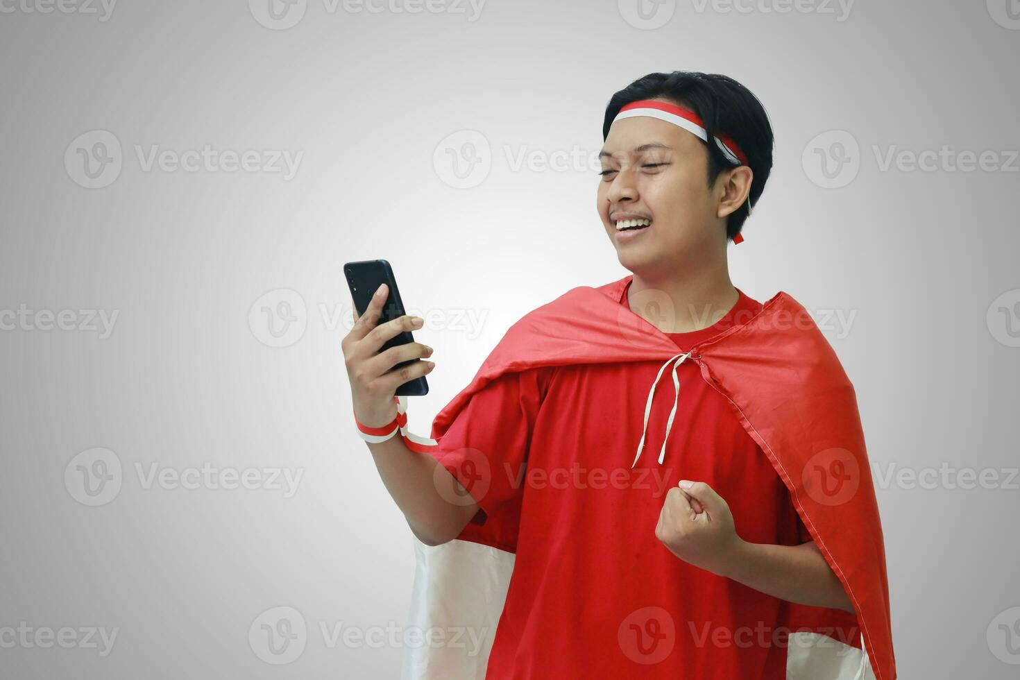 retrato de atractivo asiático hombre en camiseta con rojo blanco cinta en cabeza con bandera en su hombro como un capa, participación móvil teléfono con elevado puño. aislado imagen en gris antecedentes foto