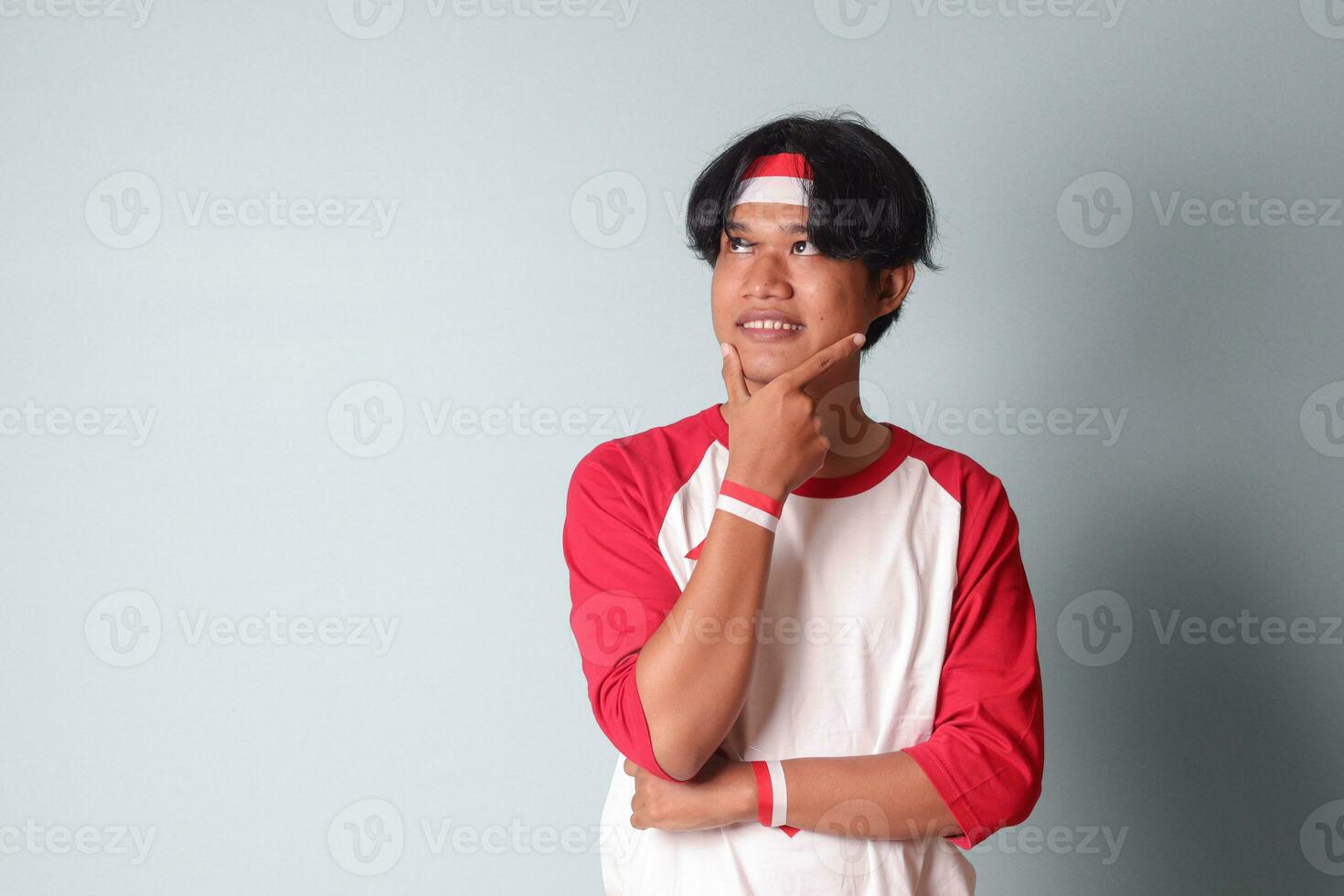Portrait of attractive Asian man in t-shirt with red and white ribbon on head, standing against gray background, thinking about question with hand on chin photo