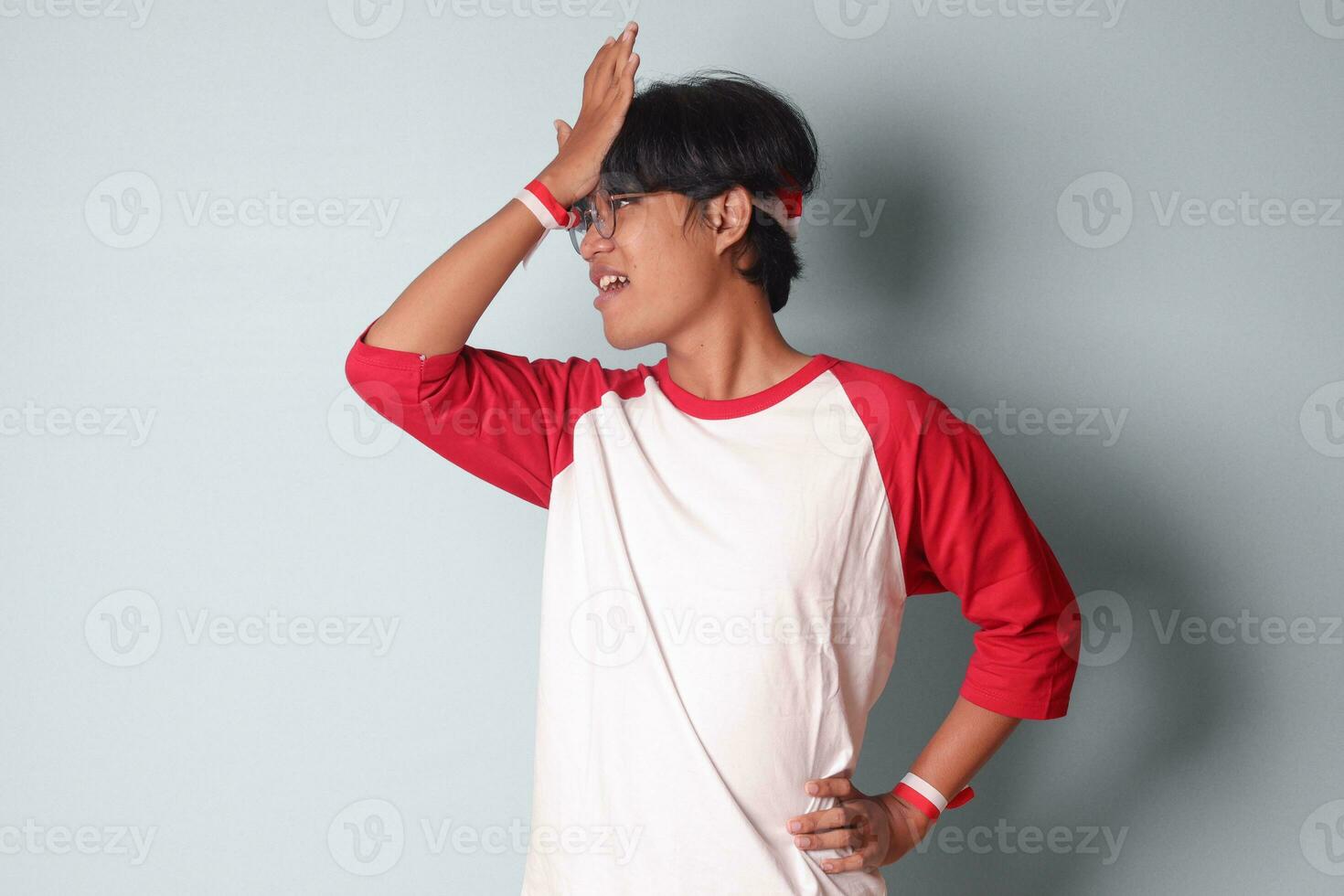 retrato de atractivo asiático hombre en camiseta con rojo y blanco cinta en cabeza, teniendo un migraña, conmovedor su templo. dolor de cabeza concepto. aislado imagen en gris antecedentes foto
