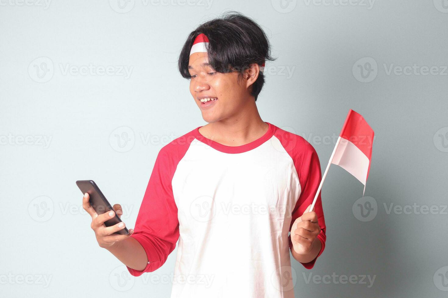 Portrait of attractive Asian man in t-shirt with red and white ribbon on head, holding mobile phone while raising up Indonesia flag. Isolated image on gray background photo