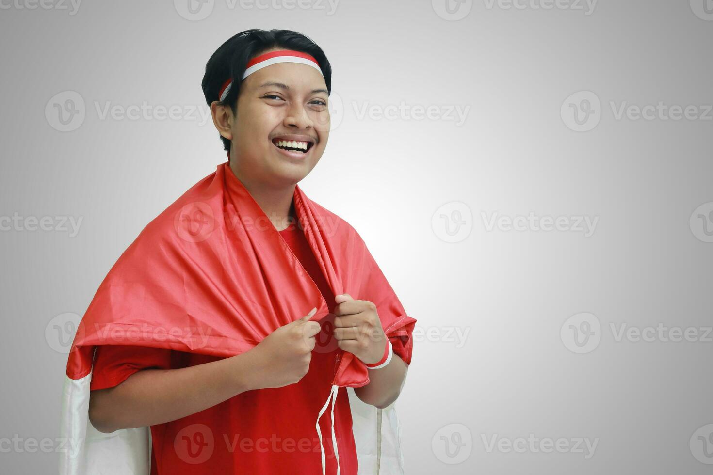 Portrait of attractive Asian man in t-shirt with red white ribbon on head with flag on his shoulder as a cloak, looking up with hands on hip. Isolated image on gray photo
