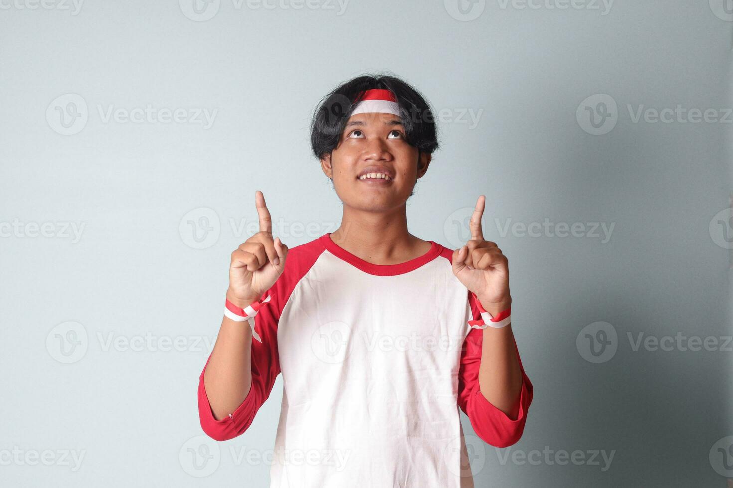 retrato de atractivo asiático hombre en camiseta con rojo y blanco cinta en cabeza, señalando a alguna cosa con dedo. aislado imagen en gris antecedentes foto