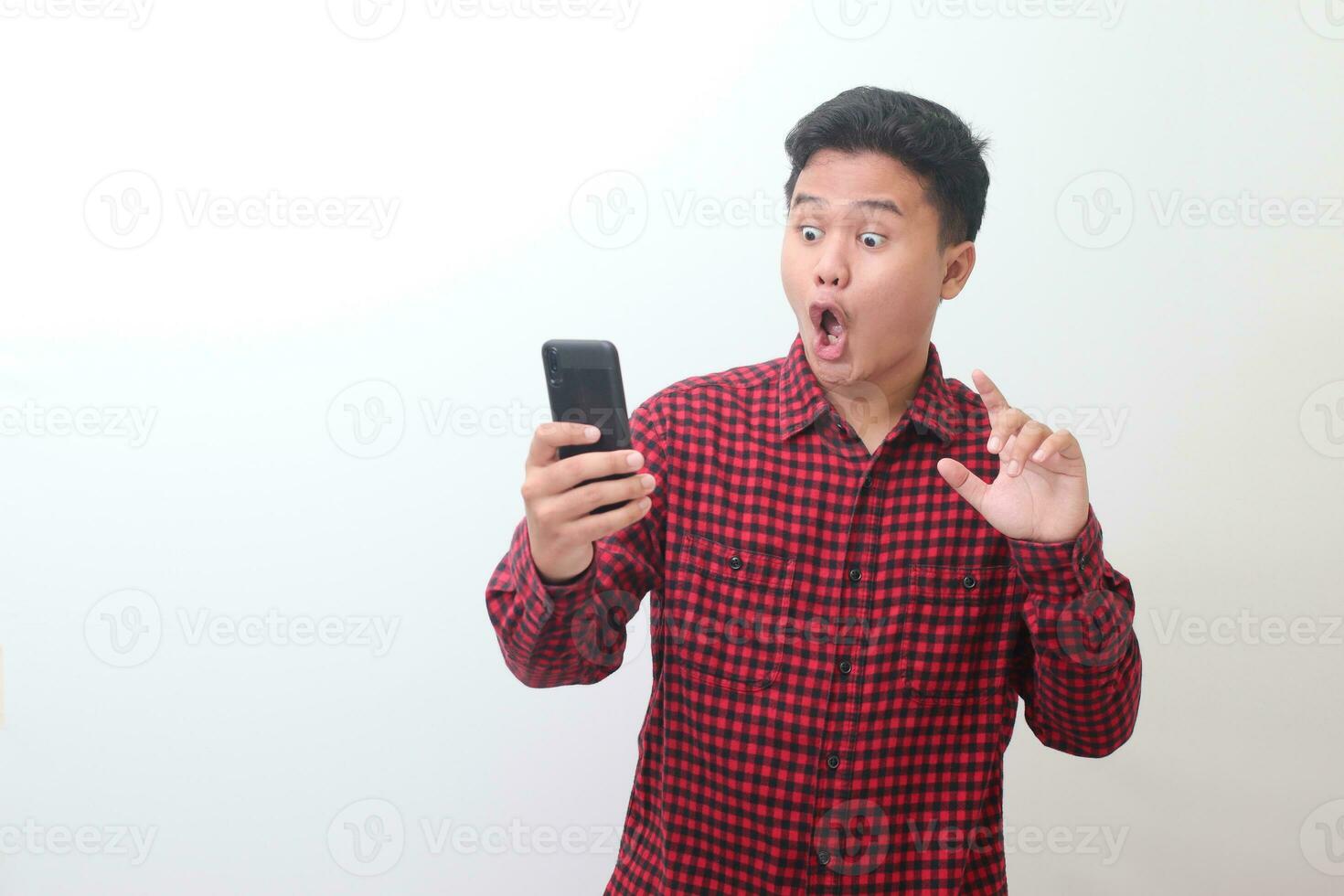 Portrait of shocked Asian man in casual plaid shirt standing against gray background, holding a mobile phone while trying to touch it. Advertising concept. photo