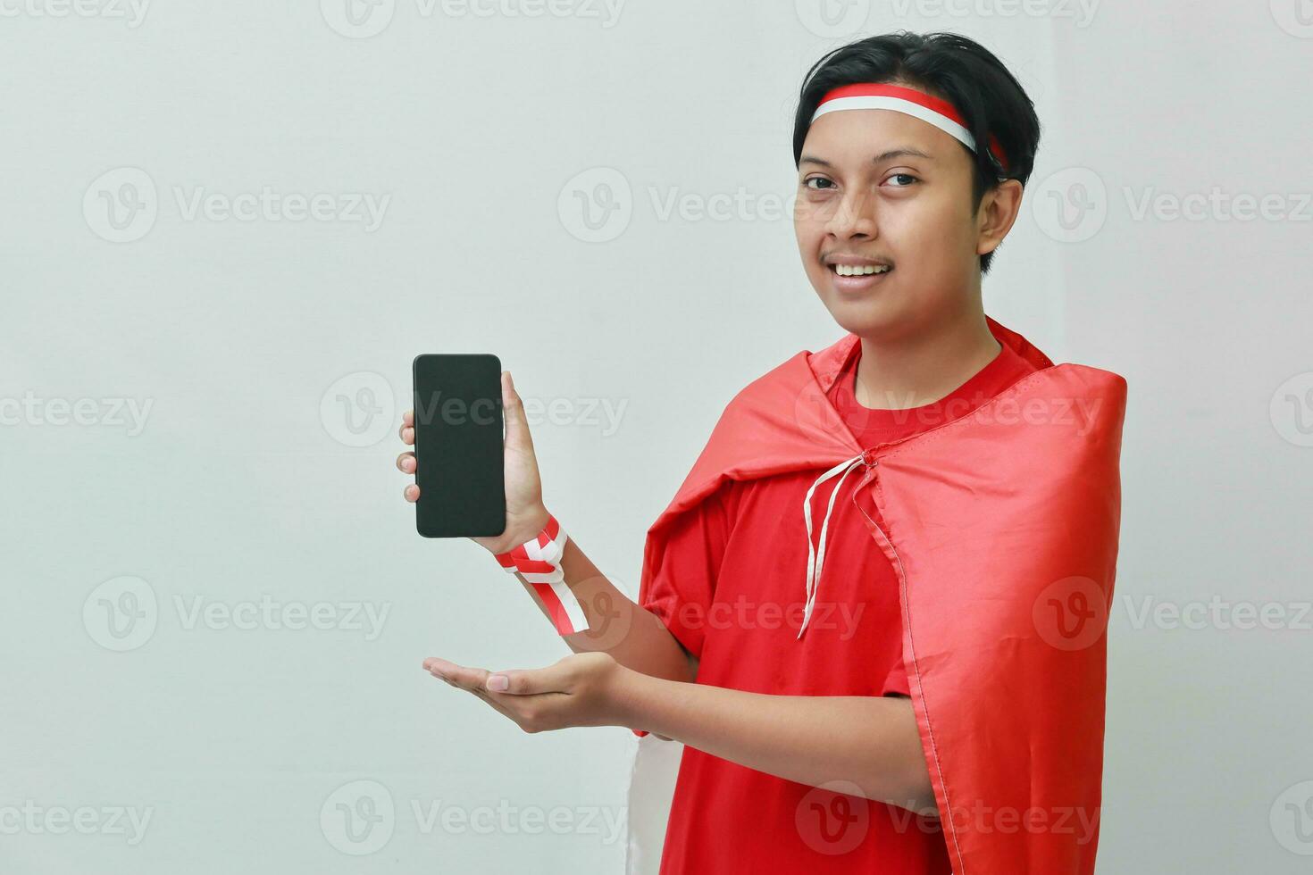 Portrait of attractive Asian man in t-shirt with red white ribbon on head with flag on his shoulder as a cloak, presenting a blank screen mobile phone. Isolated image on gray background photo