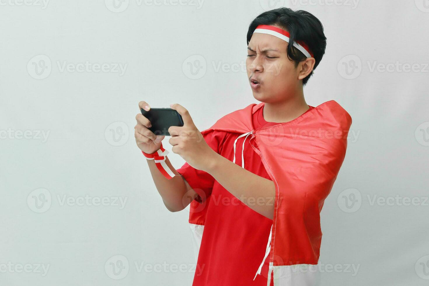 retrato de atractivo asiático hombre en camiseta con rojo blanco cinta en cabeza con bandera en su hombro como un capa, jugando juego en móvil teléfono con enojado expresión. aislado imagen en gris antecedentes foto