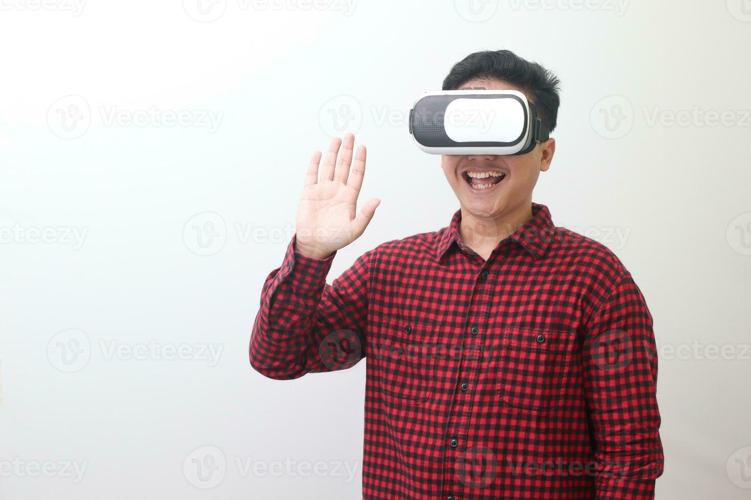 Portrait of Asian man in red plaid shirt using Virtual Reality VR glasses and greet his friend inside the game with opened palm. Isolated image on white background photo