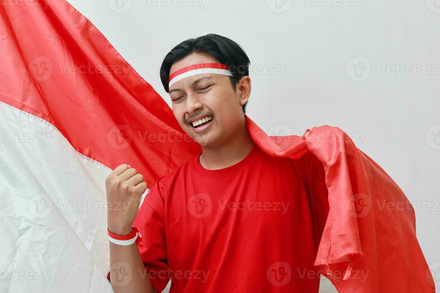 retrato de atractivo asiático hombre en camiseta con rojo y blanco cinta en cabeza, levantamiento bandera con su puño, celebrando de indonesia independencia día. aislado imagen en gris antecedentes foto