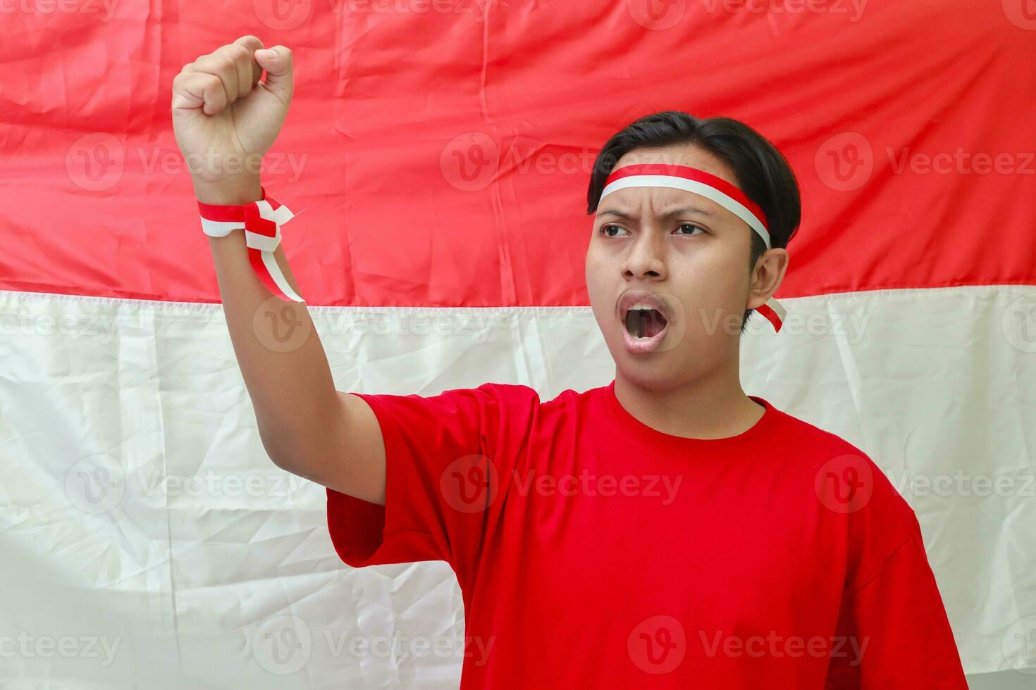 Portrait of attractive Asian man in t'shirt with red and white ribbon on head, raising up his arm and shouting. Guy standing in front of Indonesian flag photo