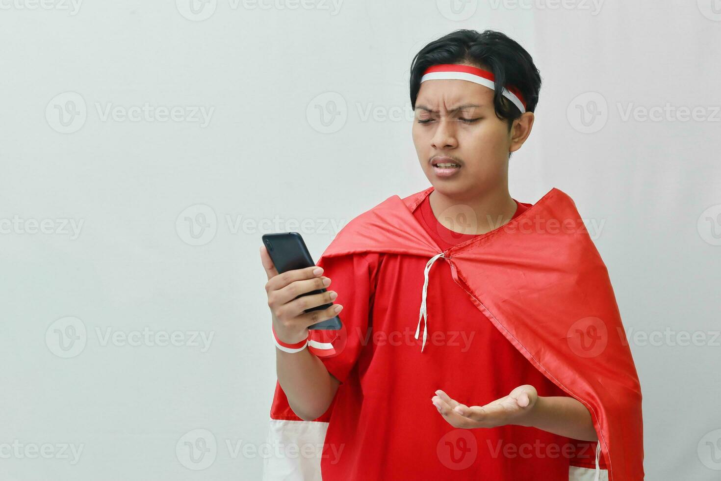 Portrait of attractive Asian man in t-shirt with red white ribbon on head with flag on his shoulder as a cloak, pointing and holding mobile phone. Isolated image on gray background photo