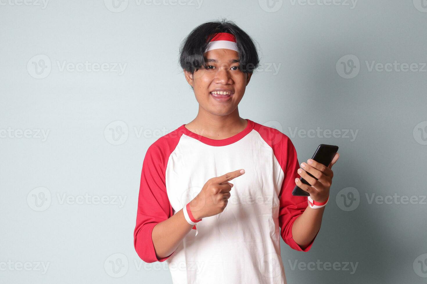 Portrait of attractive Asian man in t-shirt with red and white ribbon on head, holding mobile phone while raising up Indonesia flag and pointing away. Isolated image on gray background photo