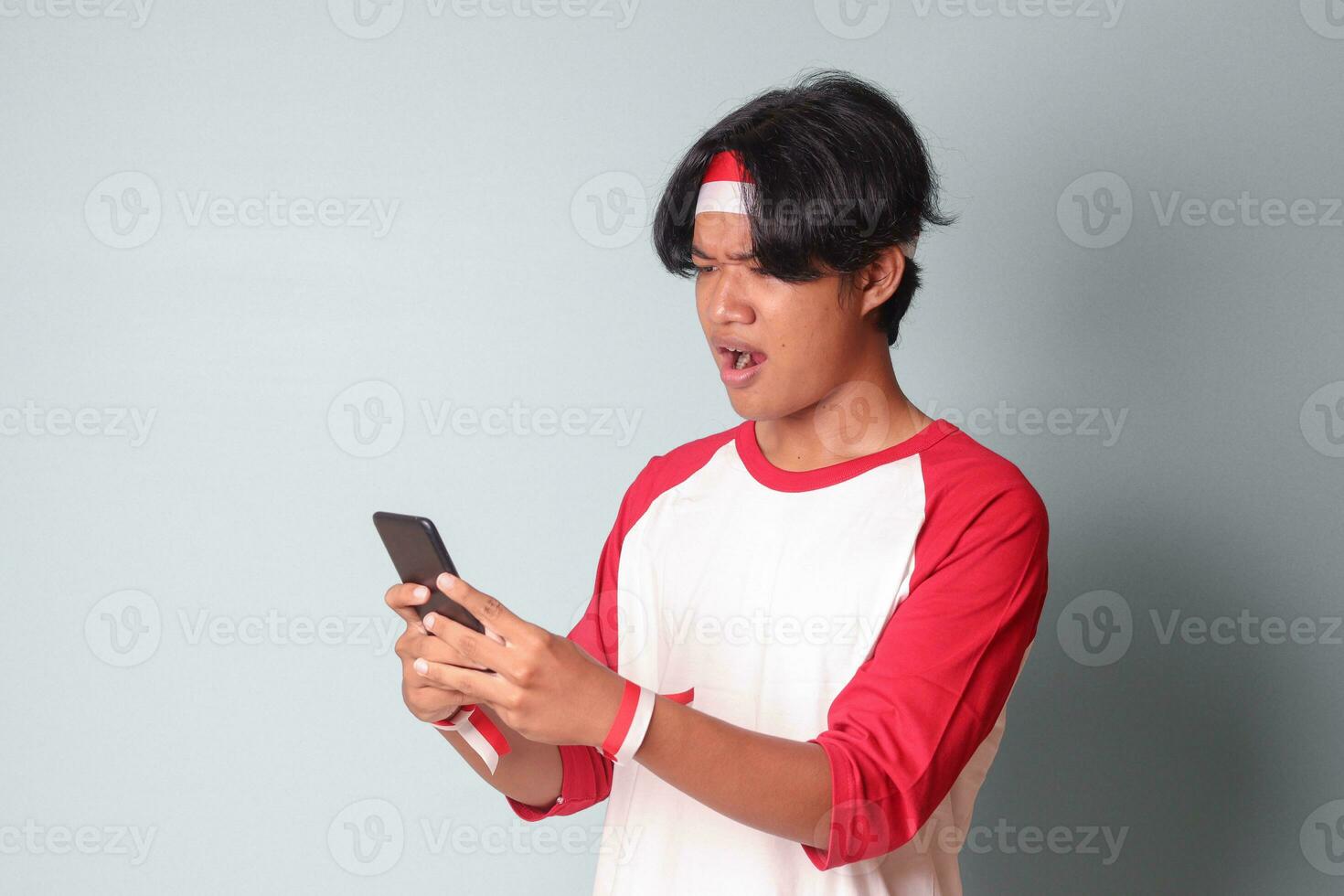 retrato de conmocionado asiático hombre en camiseta con rojo y blanco cinta en cabeza, participación móvil teléfono con sorprendido expresión. aislado imagen en gris antecedentes foto